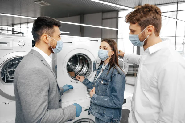 Vendedor en hipermercado con máscara médica demuestra a sus clientes una nueva lavadora — Foto de Stock