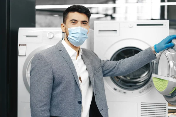 Asistente de tienda masculino en máscara médica demuestra un nuevo modelo de lavadora en el hipermercado — Foto de Stock