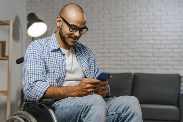 Joven afroamericano en gafas sentado en silla de ruedas y usando su teléfono inteligente — Foto de Stock