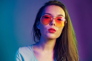 Güneş gözlüklü, siyah deri ceketli koyu mavi arka planda poz veren Asyalı kız.