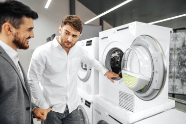 Hipermarkette gülümseyen bir satıcı müşteriyle çamaşır makinesi hakkında konuşuyor.