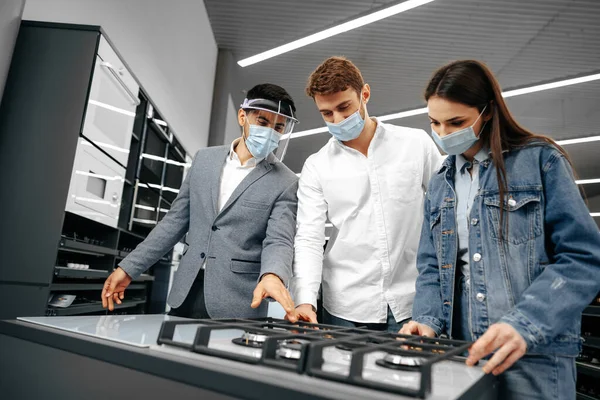 Bolti asszisztens a hipermarketben mutatja az új gáztűzhely modellt fiatal párnak, akik orvosi maszkot viselnek. — Stock Fotó