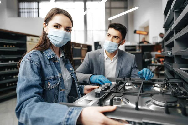 Man säljare i stormarknad beskriver funktioner i gasspis till kvinnliga kunder, båda bär ansiktsmasker — Stockfoto