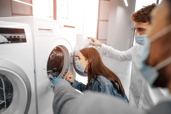 Vendedor en hipermercado con máscara médica demuestra a sus clientes una nueva lavadora — Foto de Stock