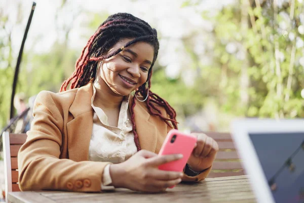 Joven mujer afroamericana sentada en la cafetería de la calle y sosteniendo el teléfono inteligente en sus manos — Foto de Stock