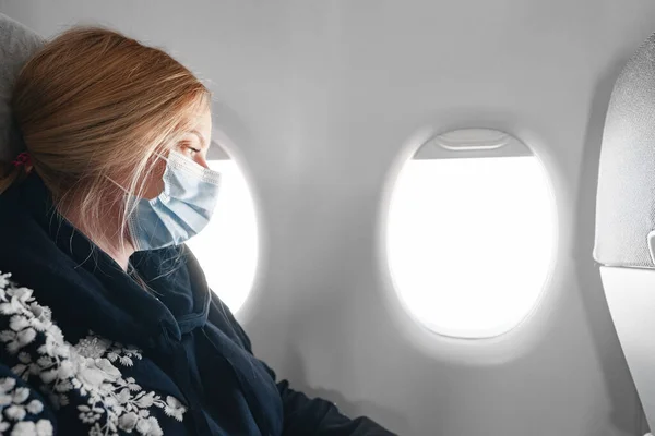 Блондинка в медицинской маске сидит в самолете у окна — стоковое фото