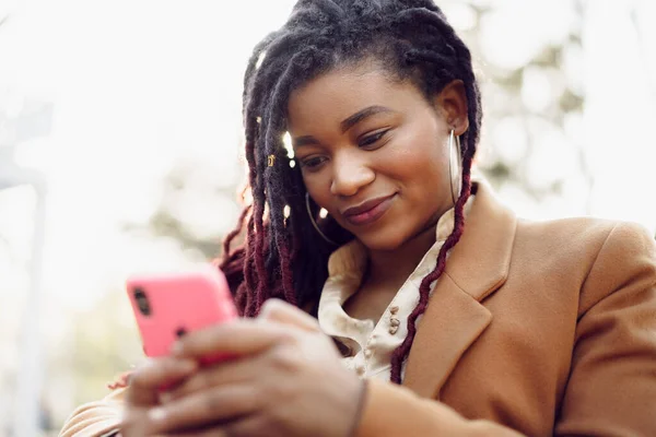 Joven mujer afroamericana sentada en la cafetería de la calle y sosteniendo el teléfono inteligente en sus manos — Foto de Stock