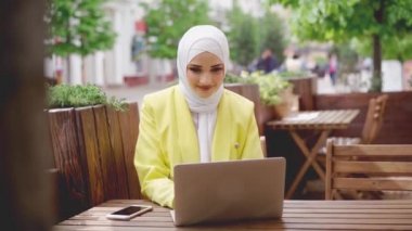 Başörtüsü takan gülümseyen genç Müslüman kadın kafede oturur ve dizüstü bilgisayar kullanır.