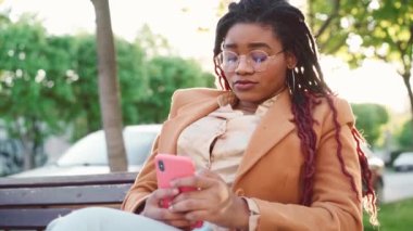 Genç Afrikalı Amerikalı kadın şehirde bir bankta oturuyor ve akıllı telefonunu kullanıyor.