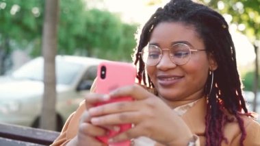 Genç Afrikalı Amerikalı kadın şehirde bir bankta oturuyor ve akıllı telefonunu kullanıyor.