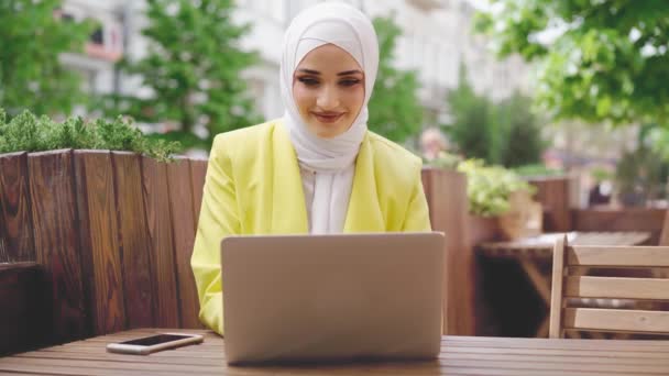 Sonriente joven musulmana mujer con pañuelo en la cabeza se sienta en la cafetería y utiliza el ordenador portátil — Vídeo de stock