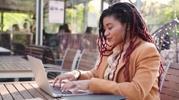 Młoda Afroamerykanka siedzi w kawiarni na świeżym powietrzu i używa laptopa — Wideo stockowe