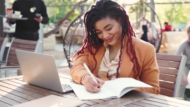 Jonge Afrikaanse Amerikaanse vrouw zit in outddor cafe met laptop en het maken van aantekeningen in notitieblok — Stockvideo
