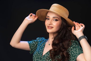 Hasır şapkalı, siyah arka planda poz veren güzel bir genç kadın.