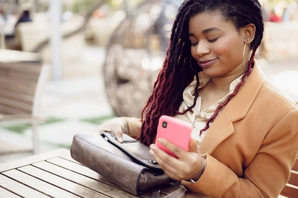 Joven mujer afroamericana sentada en la cafetería de la calle y sosteniendo el teléfono inteligente en sus manos — Foto de Stock