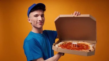 Mavi üniformalı genç teslimatçı sarı arka planda bir kutu taze pizza açıyor.