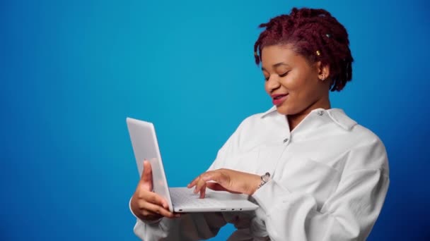 Молодая афро-женщина держит ноутбук и печатает на синем фоне — стоковое видео