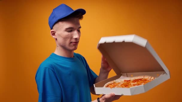 Молодой курьер в синей форме открывает коробку свежей пиццы на желтом фоне — стоковое видео