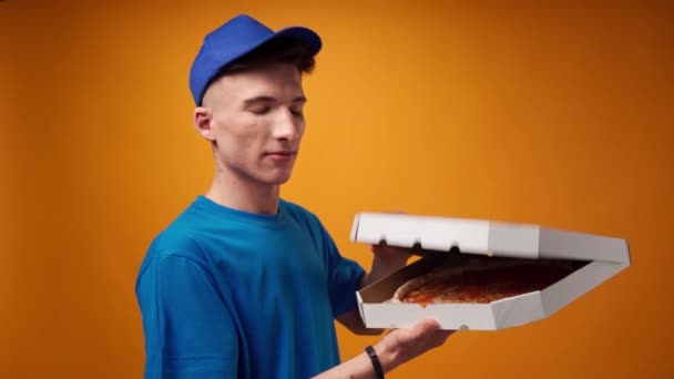 Jonge bezorger in blauw uniform opent een doos verse pizza tegen gele achtergrond — Stockvideo