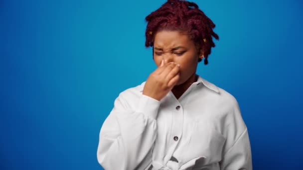 Afro vrouw in wit shirt sluit neus vanwege een slechte stinkende geur, blauwe achtergrond — Stockvideo