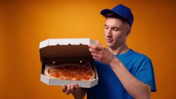 Młody dostawca w niebieskim mundurze otwiera pudełko świeżej pizzy na żółtym tle — Wideo stockowe