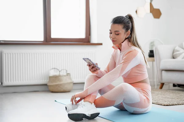 Beautiful young woman in sportswear sitting indoors and looking at smartphone before workout — Stock Photo, Image