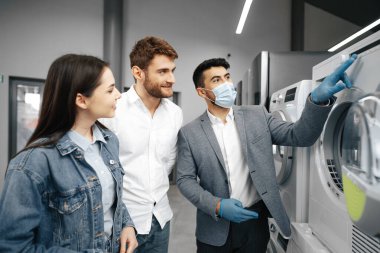 Hipermarkette tıbbi maske takan satıcı müşterilerine yeni bir çamaşır makinesi gösteriyor.
