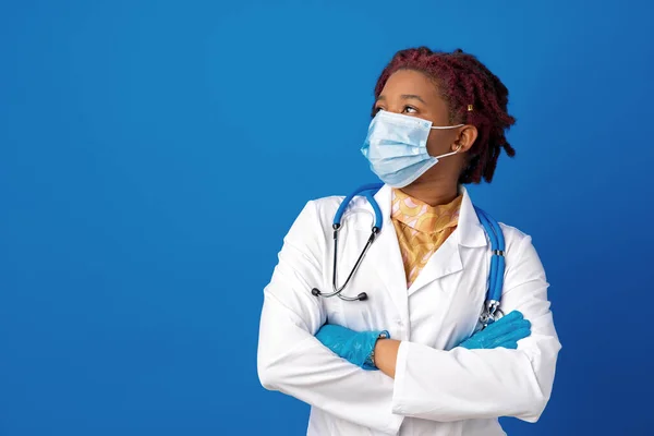 Porträt einer afrikanischen Ärztin im Laborkittel mit Gesichtsmaske und Stethoskop vor blauem Hintergrund — Stockfoto