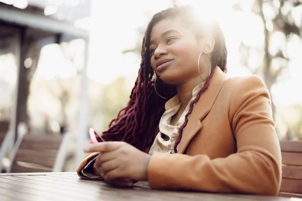 Joven mujer afroamericana sentada en la cafetería de la calle y sosteniendo el teléfono inteligente en sus manos — Foto de Stock