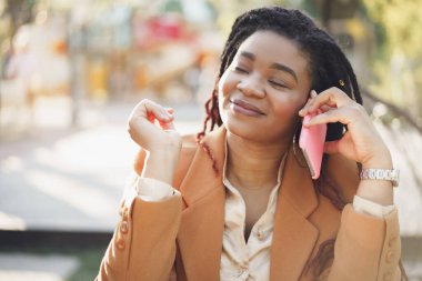 Pozitif genç siyah kadın açık kafede oturuyor ve telefonla konuşuyor.