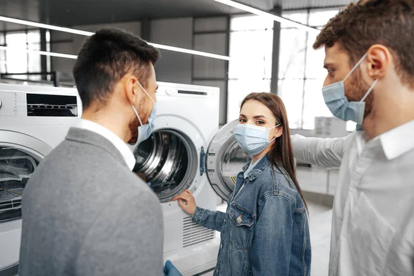 Vendedor en hipermercado con máscara médica demuestra a sus clientes una nueva lavadora — Foto de Stock