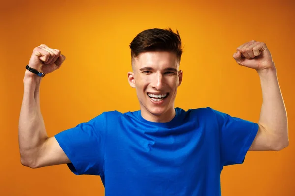 Glücklicher junger Mann im blauen T-Shirt lächelt mit erhobenen Händen und feiert den Erfolg — Stockfoto