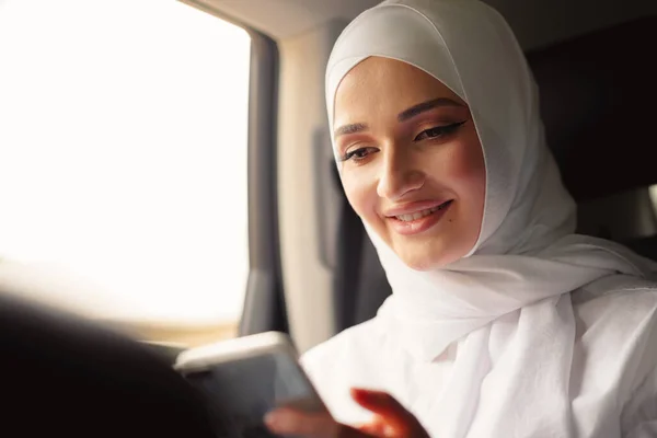 Gyönyörű muszlim nő visel fehér hidzsáb ül a hátsó ülésen egy autó és okostelefon használata — Stock Fotó