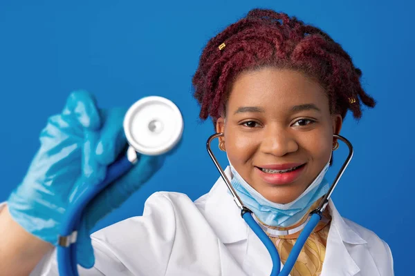 Portrait of african female doctor in lab coat with face mask and stethoscope against blue background — Stock Photo, Image