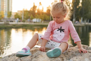 Küçük kız gölün yanındaki parkta büyük bir taşın üzerinde oturuyor.