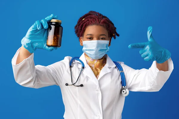 Maskierte afrikanisch-amerikanische Ärztin zeigt Glas mit Medizin vor blauem Hintergrund — Stockfoto