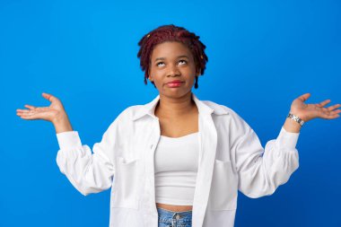 Hesitant african american woman shrugging her shoulders against blue background clipart
