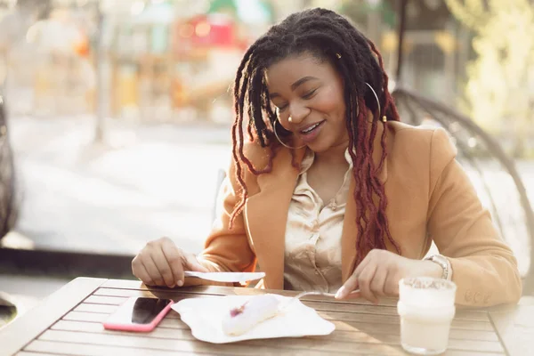 Sonriente mujer afroamericana bebiendo café y comiendo postre en la cafetería al aire libre — Foto de Stock