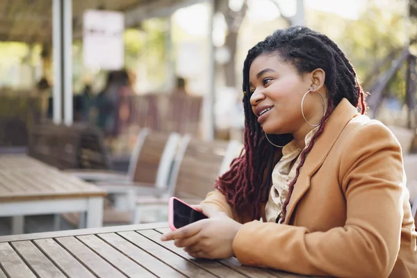 Joven mujer afroamericana sentada en la cafetería de la calle y sosteniendo el teléfono inteligente en sus manos — Foto de Stock