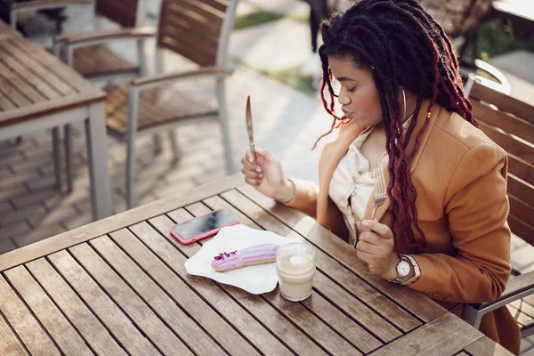 Sonriente mujer afroamericana bebiendo café y comiendo postre en la cafetería al aire libre — Foto de Stock