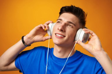 Mavi tişörtlü genç adam sarı arka planda kulaklıkla müzik dinliyor.