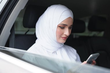 Beyaz tesettürlü güzel Müslüman kadın arabanın arka koltuğunda oturuyor ve akıllı telefon kullanıyor.