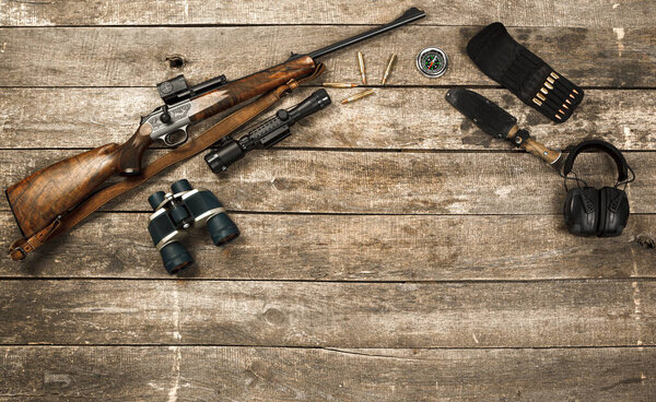 Hunting equipment on old wooden background including rifle, knife, binoculars and cartridges
