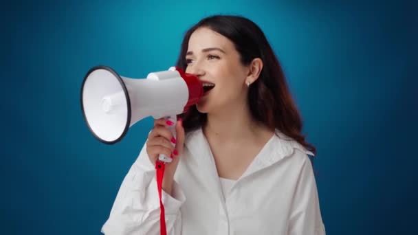 Mooie jonge vrouw schreeuwen in luidspreker tegen blauwe achtergrond — Stockvideo