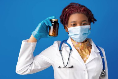 Maskeli Afrikalı Amerikalı kadın doktor mavi arka planda cam ilaç kavanozu gösteriyor.