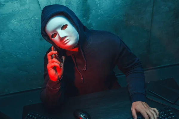 Anonymous hacker wearing face mask working on computer in dark room
