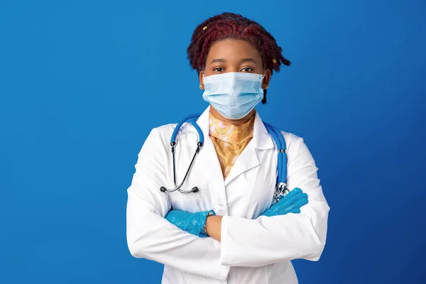 Porträt einer afrikanischen Ärztin im Laborkittel mit Gesichtsmaske und Stethoskop vor blauem Hintergrund — Stockfoto