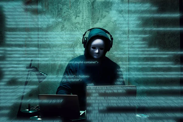 Anonymous hacker wearing face mask working on computer in dark room