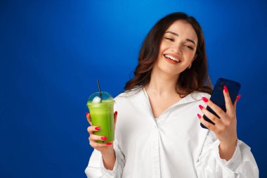 Genç, çekici, mutlu bir kadın elinde bir fincan yeşil smoothie tutuyor ve mavi arka planda akıllı telefonunu kullanıyor.