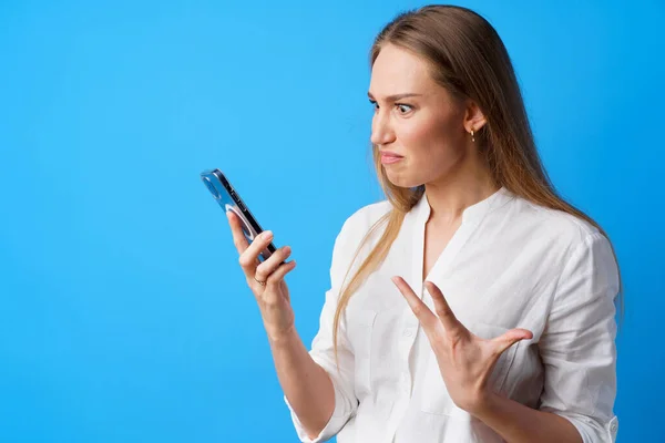 Portrait of young angry woman using her phone, annoyed texting with someone, blue background — Stock Photo, Image